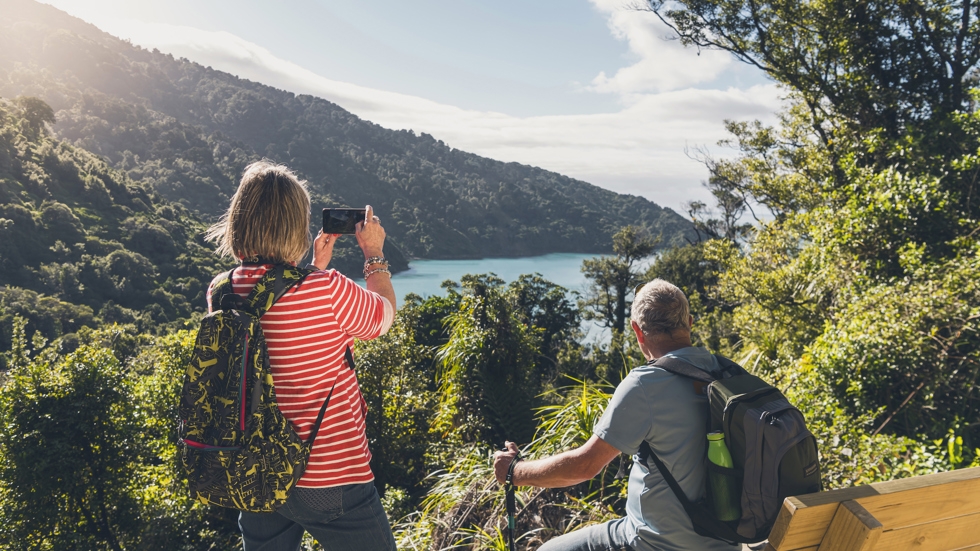 A woman takes a photo on her phone of the view across Ship Cove/Meretoto while a man sits on a bench on the northern Queen Charlotte Track in the Marlborough Sounds, New Zealand.