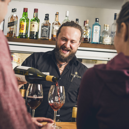 A friendly bartender pours Pinot Noir into glasses for customers at the Boatshed Cafe and Bar at Punga Cove - choose from a great range of local craft beer, wine , cocktails, soft drinks and expresso coffee.