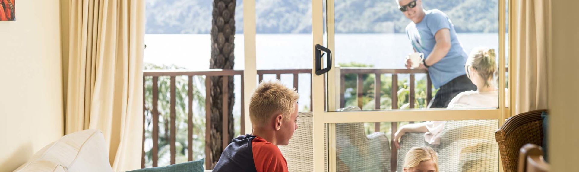 A family relaxes in the Punga Cove Frond Suites while the children play cards in the living area and adults sit on the private balcony in the Marlborough Sounds in New Zealand's top of the South Island