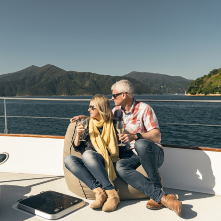 Couple sit at the bow onboard a Marlborough Tour Company boat in the Marlborough Sounds at the top of New Zealand's South Island with glasses of wine looking at the view