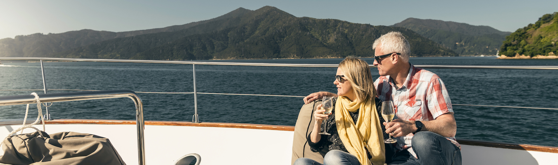 Couple sit at the bow onboard a Marlborough Tour Company boat in the Marlborough Sounds at the top of New Zealand's South Island with glasses of wine looking at the view