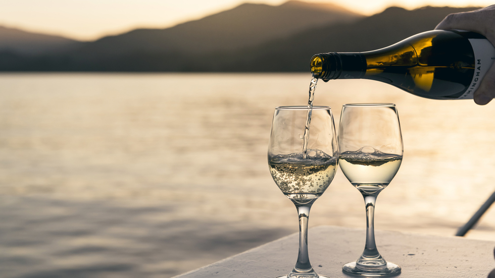 Wine bottle pouring white wine into two glasses at sunset onboard a boat in the Marlborough Sounds, at the top of New Zealand's South Island.