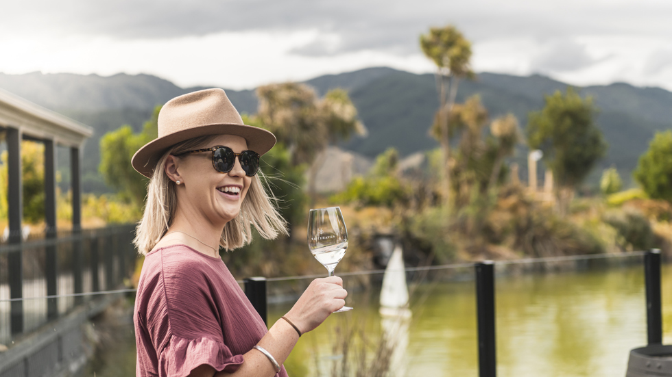 Woman with glass of wine outside in front of a pond with hill in background on a wine tour in Marlborough, at the top of New Zealand's South Island